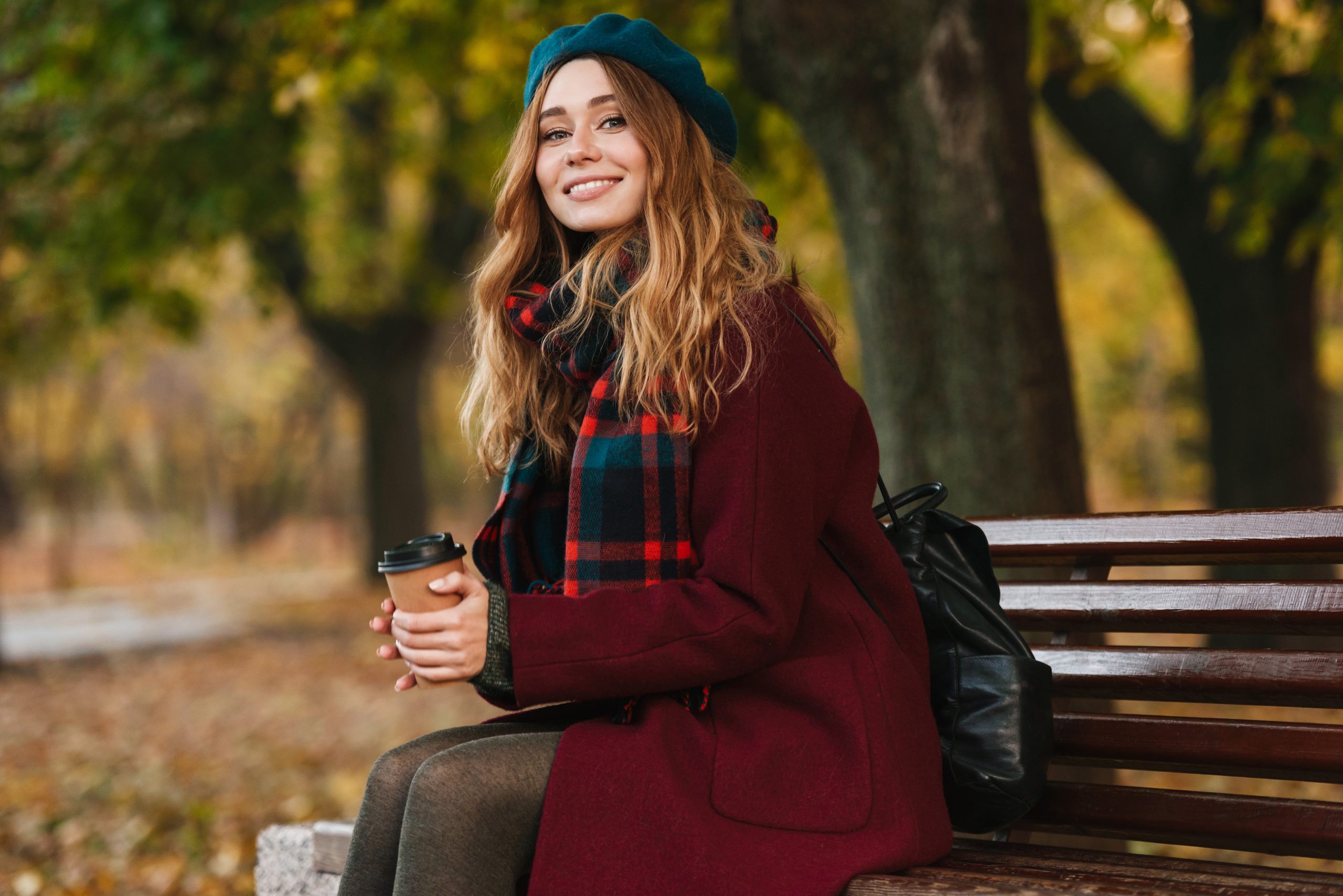 Woman wearing coat and beret sitting on bench with coffee